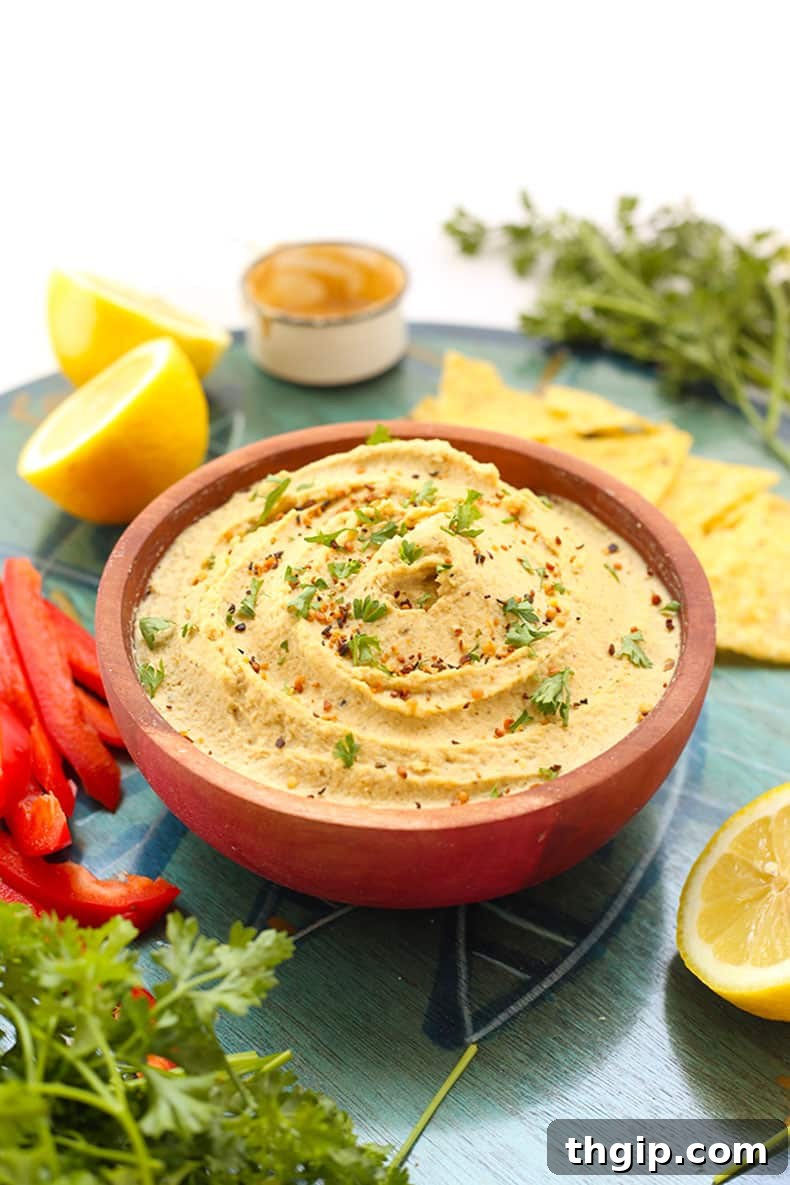 Zesty Lemon Hemp Hummus 2 Wooden bowl of bright lemon hemp hummus surrounded by fresh lemons, crispy pita chips, aromatic herbs, and finely chopped red bell peppers, ready for serving.