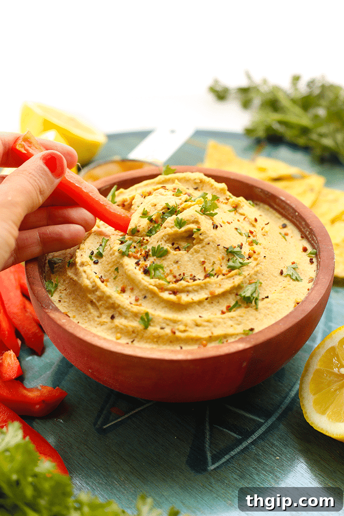 Zesty Lemon Hemp Hummus 5 Close-up shot of a hand using a slice of vibrant red bell pepper to scoop out a generous portion of creamy lemon hemp hummus from a bowl, highlighting its perfect texture and serving suggestion.