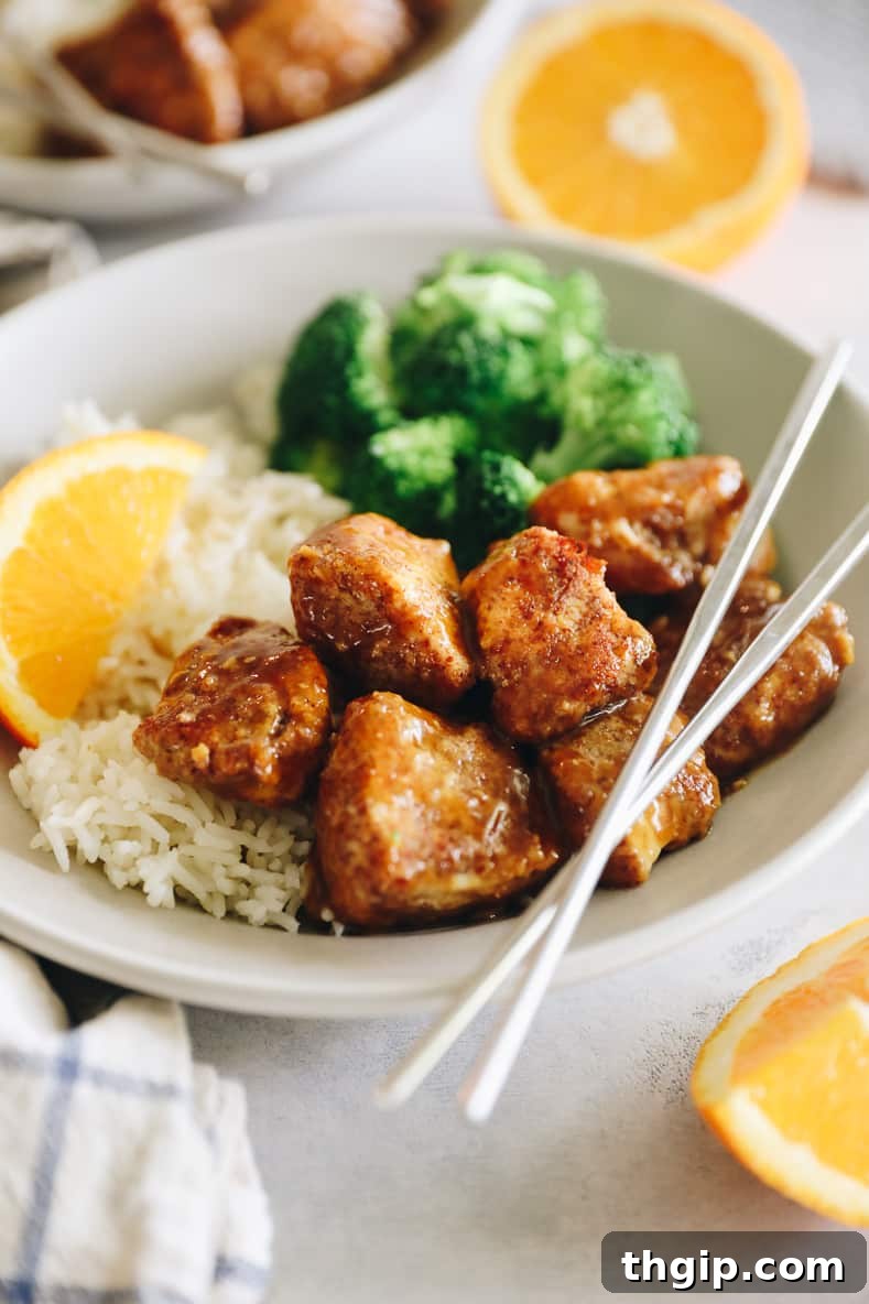 Caramelized Orange Chicken 2 Angled shot of sticky orange chicken on a white plate with rice and broccoli. Silver chopsticks crossed on top.