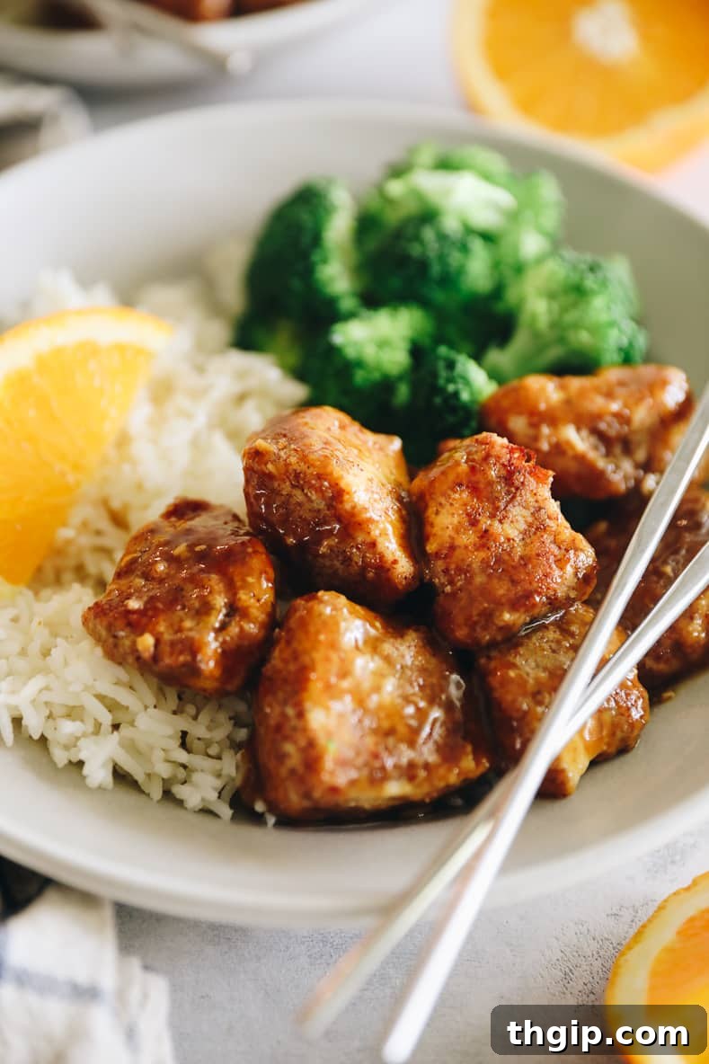 Caramelized Orange Chicken 8 Angled shot of sticky orange chicken on a white plate with rice and broccoli. Silver chopsticks crossed on top.