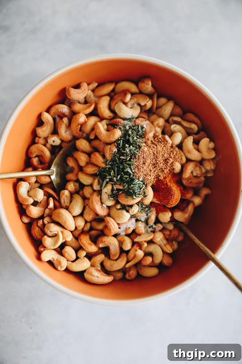 Artisan Rosemary Cashews 5 An orange bowl with cashews, rosemary and additional spices in it. Two spoons are placed in the bowl for mixing.