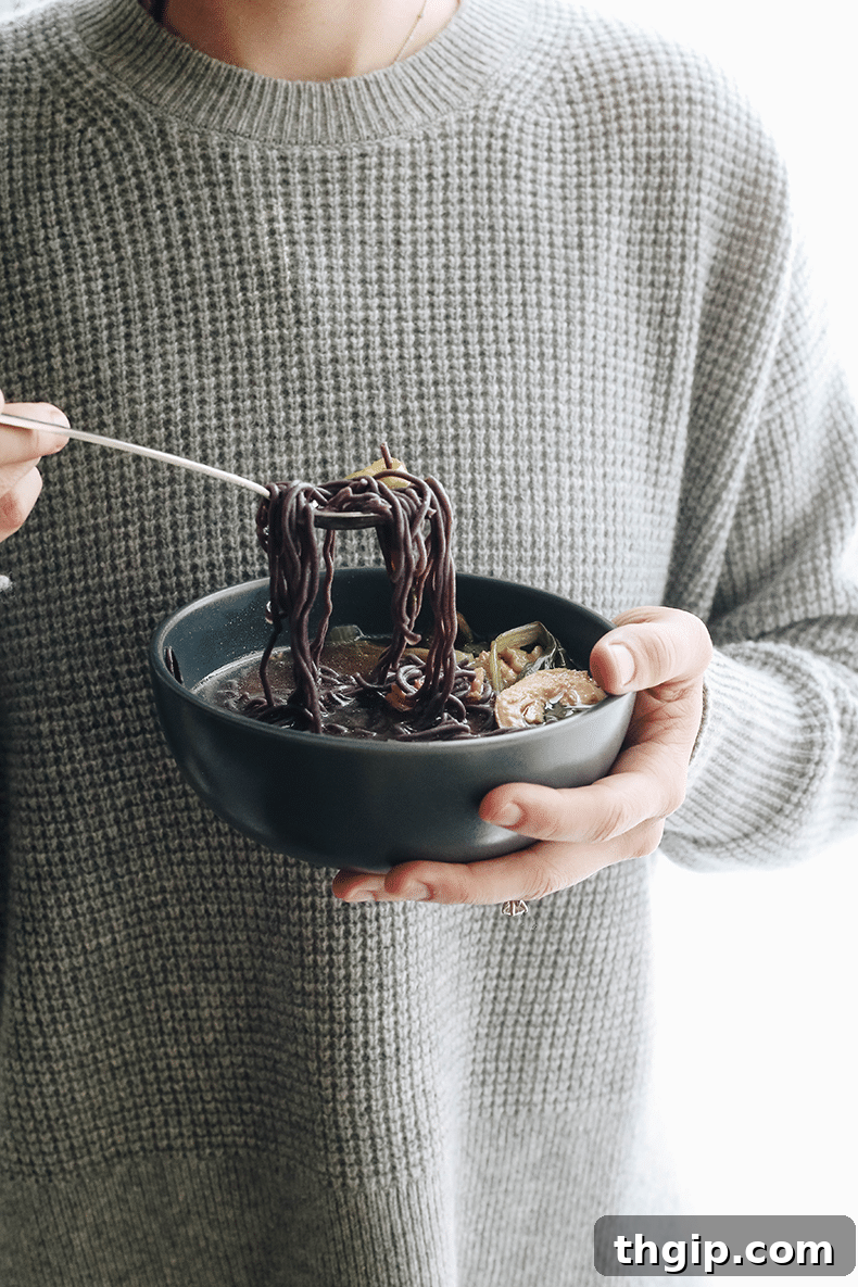 My Rejuvenating Home Remedy Bowl 3 Person in a grey waffle sweater holding a bowl of healing soup. Silver spoon taking a scoop of noodles and vegetables out of the soup.