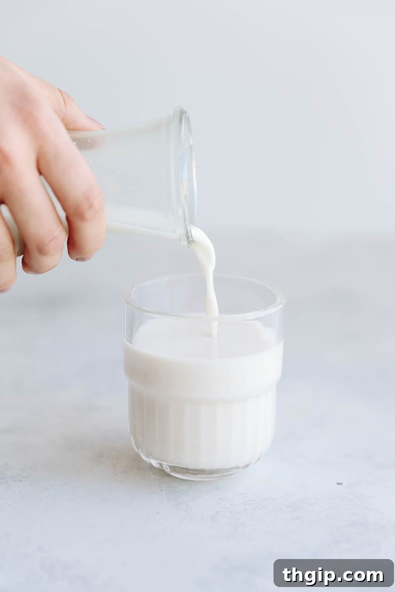 Make Your Own Oat Milk 2 Hand pouring homemade oat milk from a glass jar into a glass cup, highlighting its creamy texture.