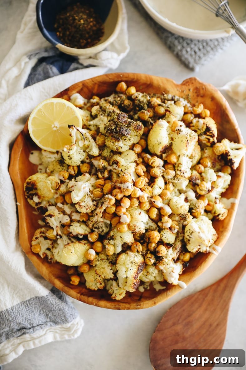 Mediterranean Roasted Cauliflower and Chickpeas with Tahini Za'atar 2 A wooden serving bowl filled with perfectly roasted cauliflower and crispy chickpeas. A wooden spoon, a small bowl of za'atar spice, and a rich tahini sauce are arranged around the main dish.