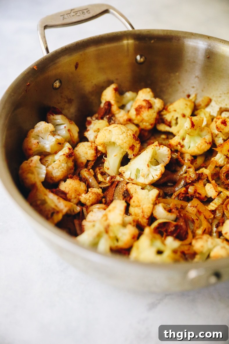 Cauliflower florets are coated with red curry paste and spices in a pan, stirring to distribute the flavors evenly.