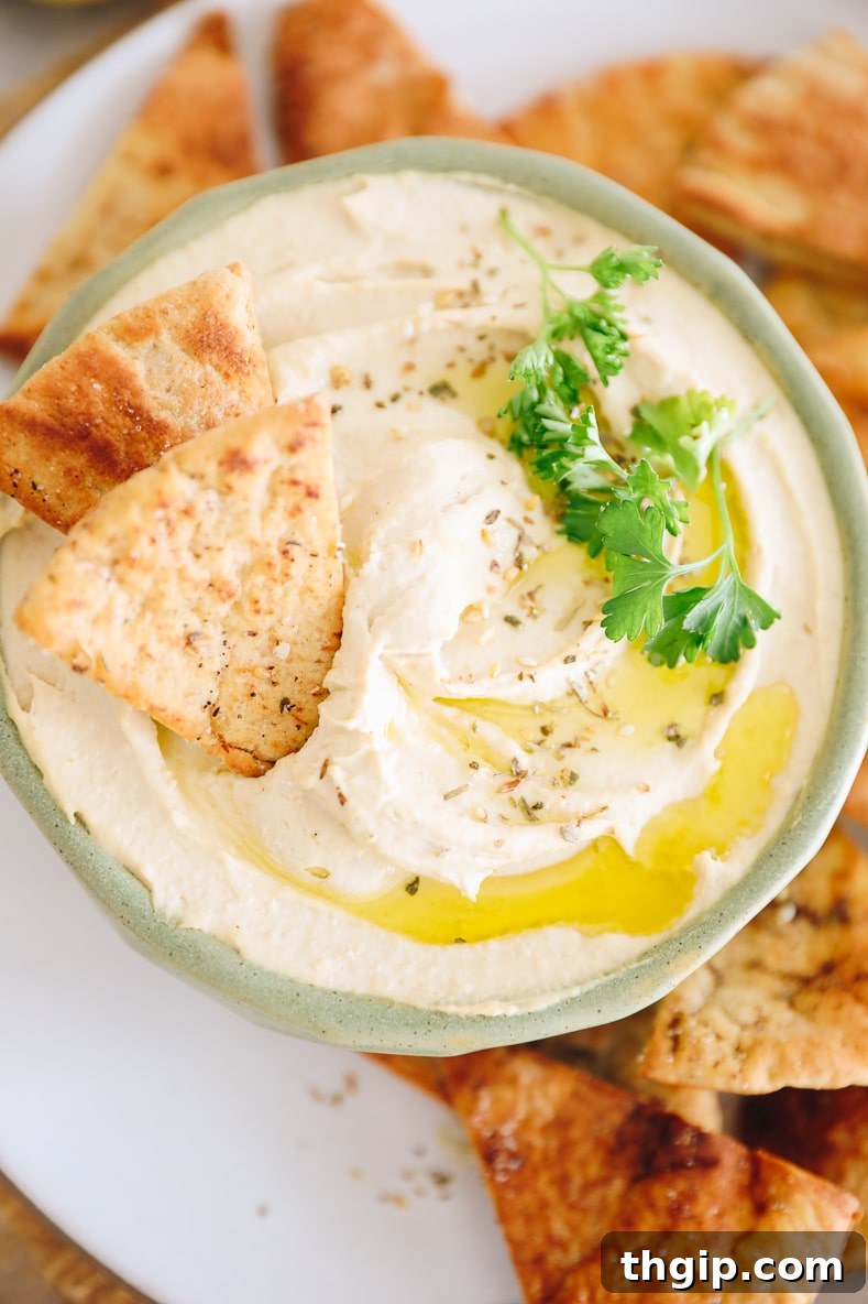 Dreamy Creamy Hummus 9 An inviting overhead shot of perfectly creamy homemade hummus in a green bowl, elegantly topped with olive oil, za'atar, and fresh parsley, surrounded by an abundance of crispy pita chips.