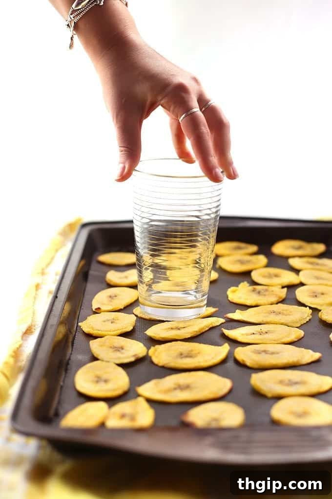 Sliced plantains being flattened on a baking sheet with the bottom of a glass.