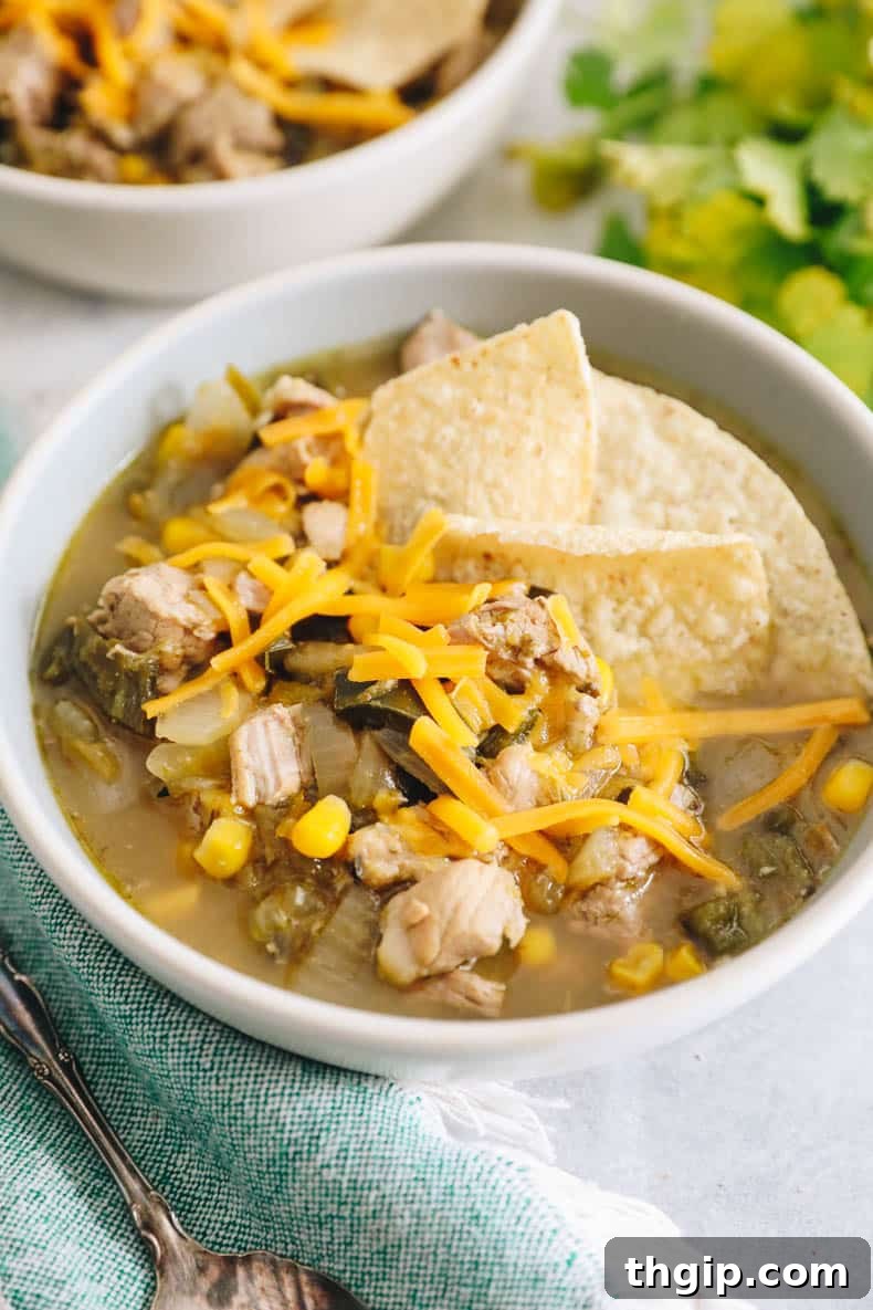 A bowl of spicy pork green chili, generously topped with shredded cheddar cheese, fresh lime wedges, and crispy tortilla chips, ready to be enjoyed on a chilly evening.