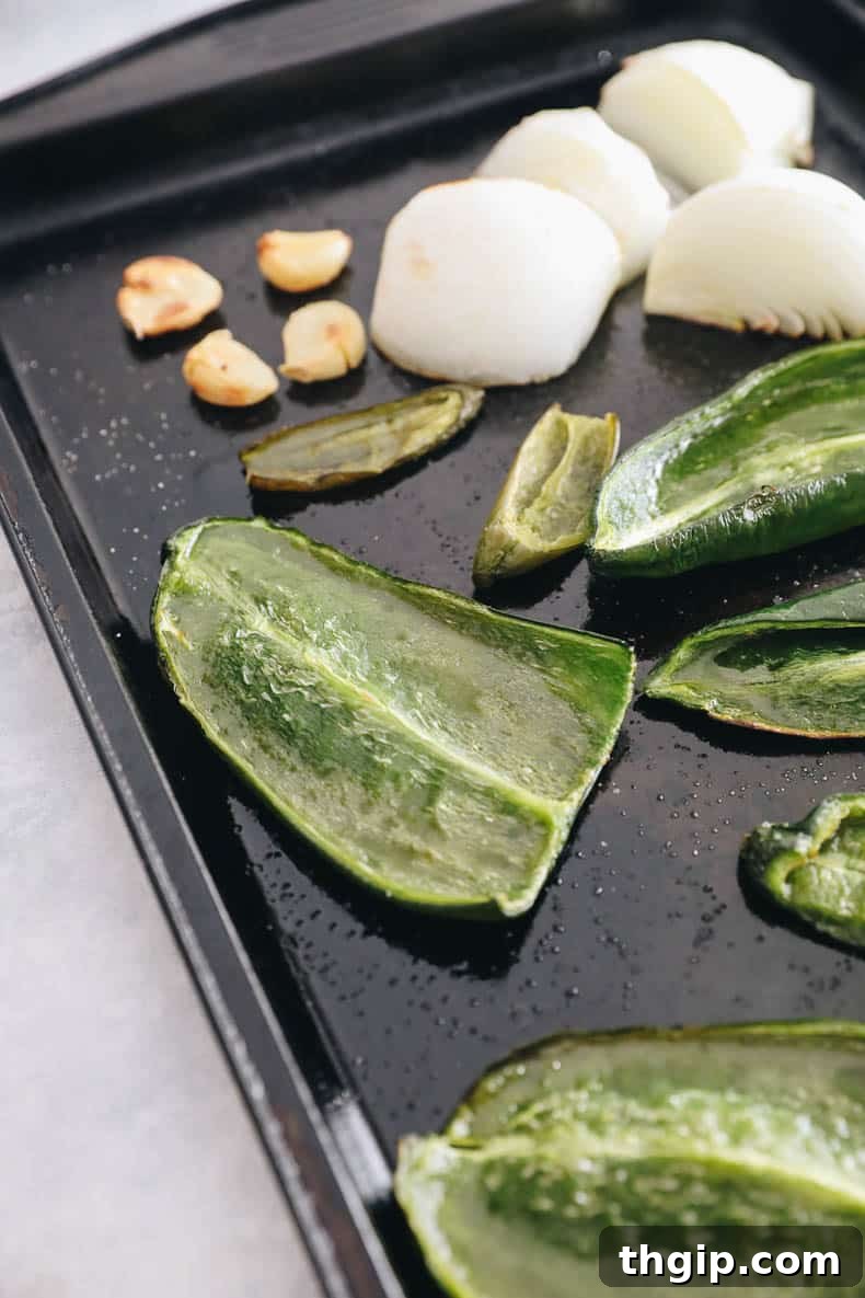 Sheet pan filled with roasted poblano peppers, jalapeno pepper, whole garlic cloves, and white onion quarters, glistening with olive oil and a sprinkle of salt.