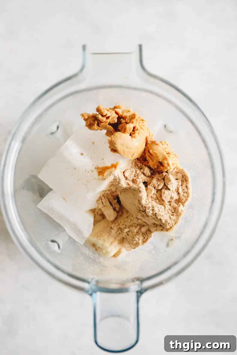 Decadent Healthy Chocolate Peanut Butter Blend 3 An overhead view inside a blender, showcasing the key ingredients for a healthy chocolate peanut butter smoothie: frozen banana slices, almond milk, creamy peanut butter, chocolate protein powder, and ice cubes, ready for blending.