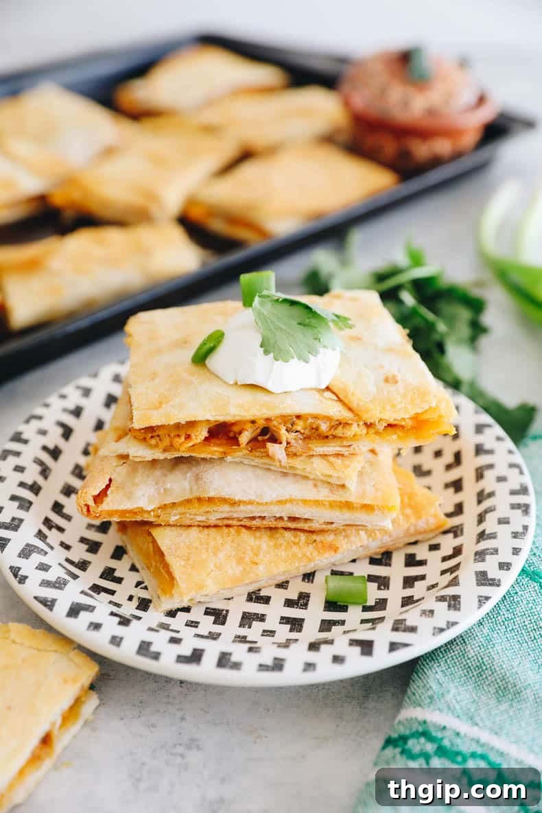 Ultimate Sheet Pan Quesadilla 2 Three golden-brown chicken quesadillas stacked on a plate, with a full sheet pan of baked quesadillas in the background, ready to be cut.