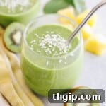 A close-up of a kiwi smoothie in a glass on a white table with a metal straw and sprinkled with coconut flakes, highlighting its fresh appeal.
