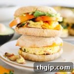 Two meal prep breakfast sandwiches inside english muffins on a white plate with avocado, tomato and cheese.