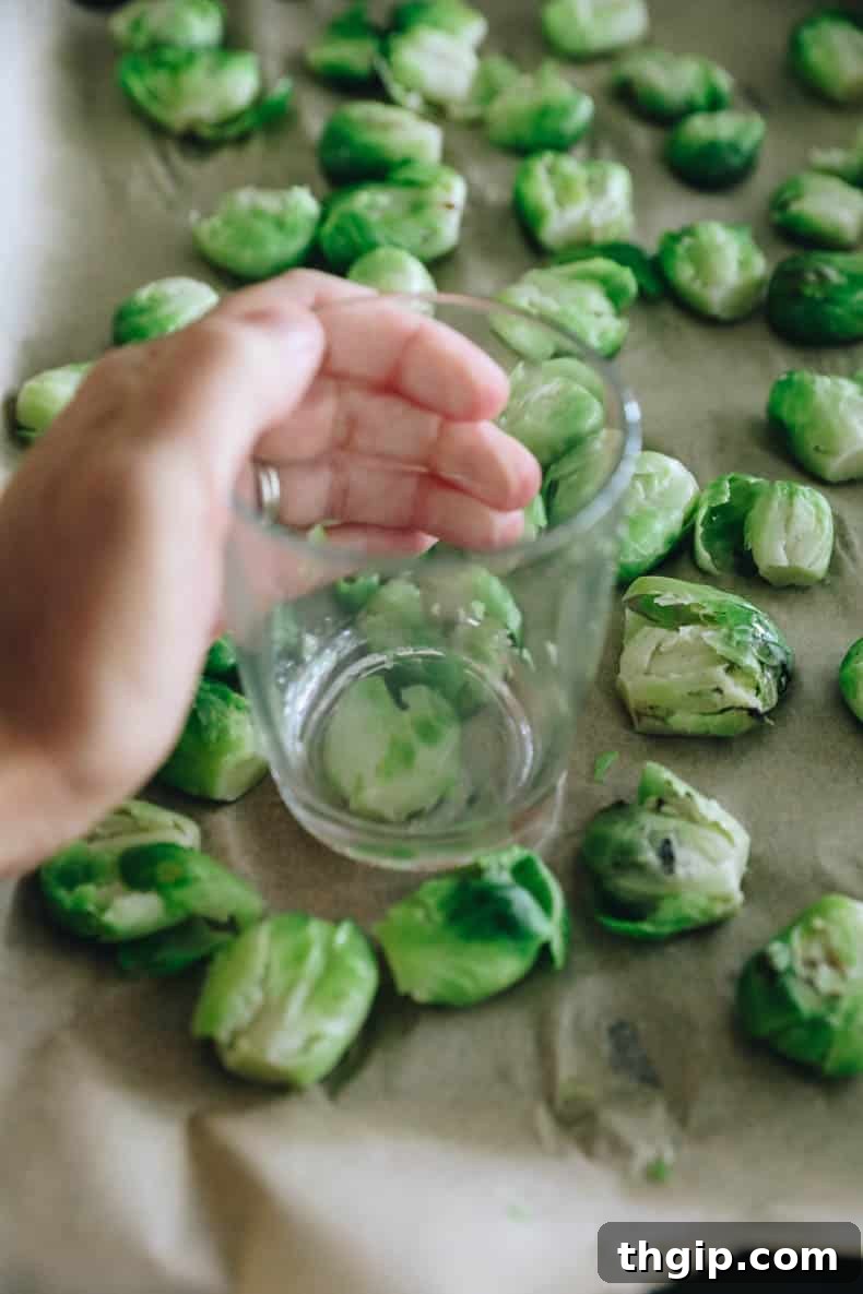 Golden Smashed Brussels Sprouts 4 Brussel sprouts being smashed with a glass cup on a parchment-lined sheet pan.