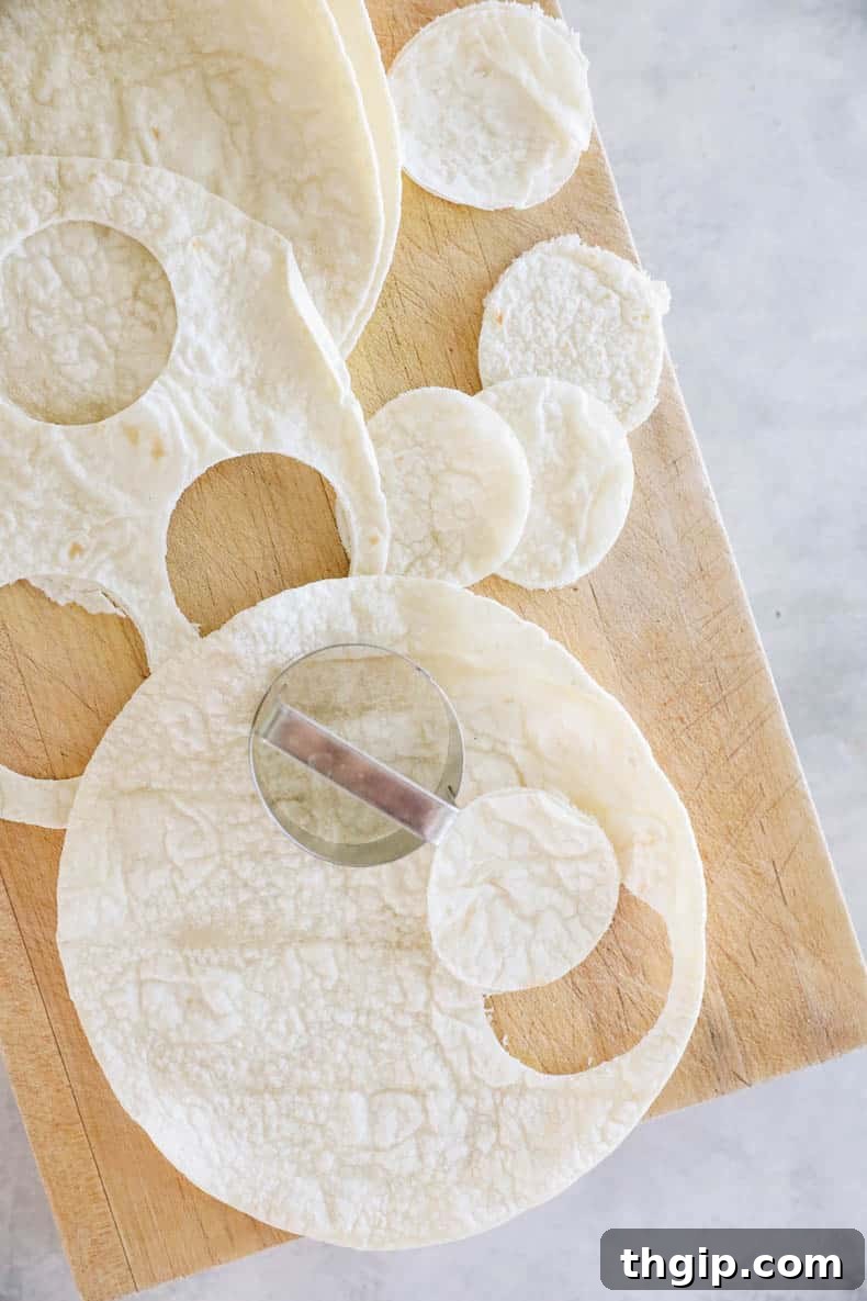 A hand using a round cookie cutter to precisely cut small circles from a large tortilla, preparing the base for mini pizza cups.