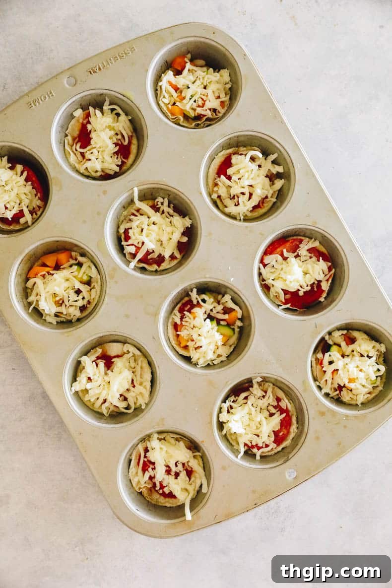 Mini pizzas being assembled in a muffin tin, each tortilla cup filled with marinara sauce, various colorful toppings, and topped with shredded mozzarella cheese, ready for baking.