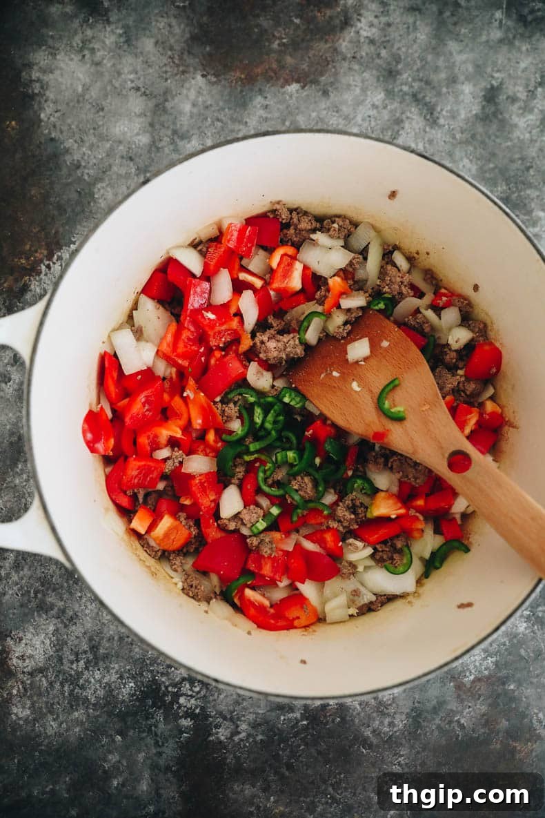 Texas-Style No-Bean Chili 4 A vibrant mix of browned beef and colorful sautéed vegetables, including onions, bell peppers, and jalapeños, seasoned with spices in a white Dutch oven.