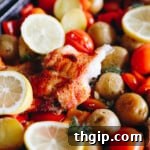 Flaky baked cod with paprika on a sheet pan, adorned with lemon slices, roasted potatoes, cherry tomatoes, and bell peppers.